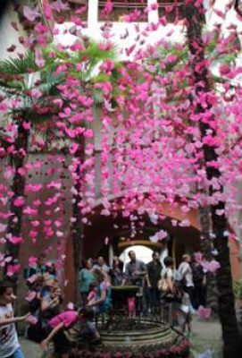 Un grupo de personas está reunida bajo un arco decorativo de arquitectura efímera adornado con flores rosas, y algunos individuos interactúan con los pétalos que caen.