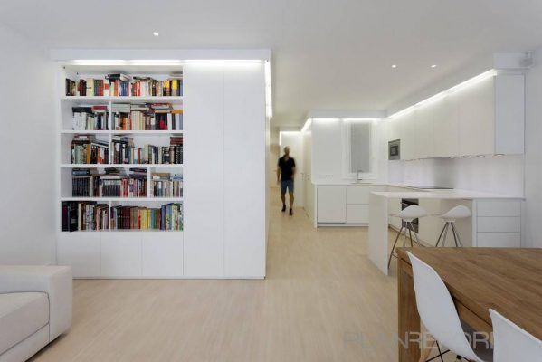Una habitación moderna y luminosa con una cocina minimalista blanca, reformas en casa como primer paso y una estantería empotrada llena de libros. Hay una silla