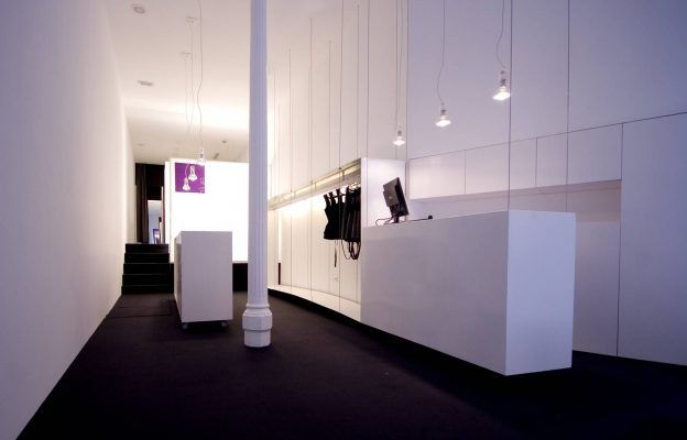 Un área de recepción moderna en una tienda de ropa contract del Proyecto inspirada en Malasaña, con un escritorio blanco, luces colgantes minimalistas y un piso oscuro, que muestra un diseño limpio y contemporáneo.