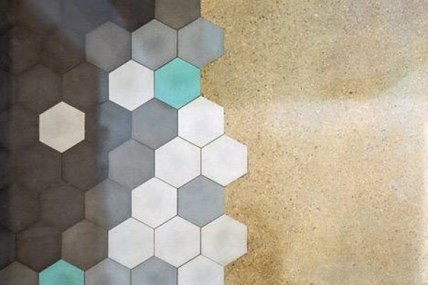 Una pared texturizada con un degradado de azulejos hexagonales que pasan del gris oscuro al gris claro, incorporando toques de amarillo y en una superficie de concreto simple.