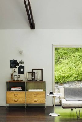 Sala de estar moderna con un aparador de madera vintage, elementos decorativos y un sofá blanco frente a un gran ventanal con vista al follaje verde, perfecto para disfrutar tu estadía en la cabaña.