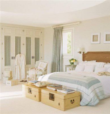 Un dormitorio cuidadosamente organizado, decorado piso por un interiorista, con una cama grande, adornada con ropa de cama blanca y azul claro, flanqueada por mesas auxiliares estilo equipaje y un cómodo sillón.
