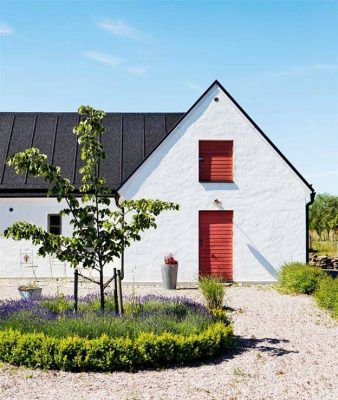Una casa blanca con contraventanas rojas bajo un cielo azul claro, de estilo nórdico, rodeada por un jardín con lavanda y un camino de grava.