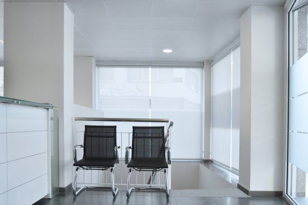 Una moderna sala de espera de oficina escasamente amueblada con dos sillas negras frente a una mesa con tapa de vidrio, bajo iluminación fluorescente blanca, rediseñada por Arquitectura Produccions siguiendo una reforma de oficina.