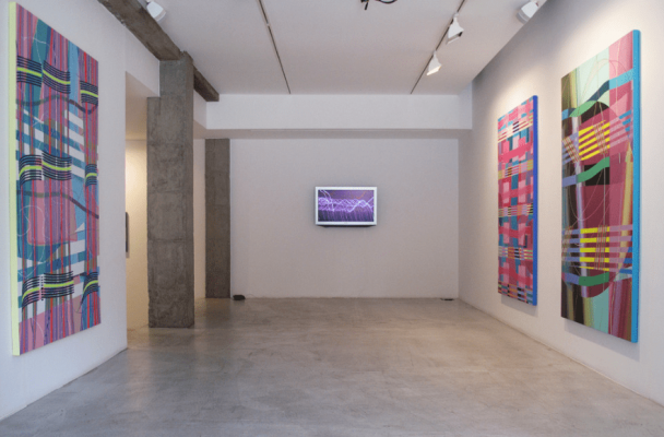 El interior de una galería de arte muestra coloridas pinturas abstractas y una exhibición de arte digital en una pared blanca, que presenta el proyecto fotográfico de Teresa Lanceta.