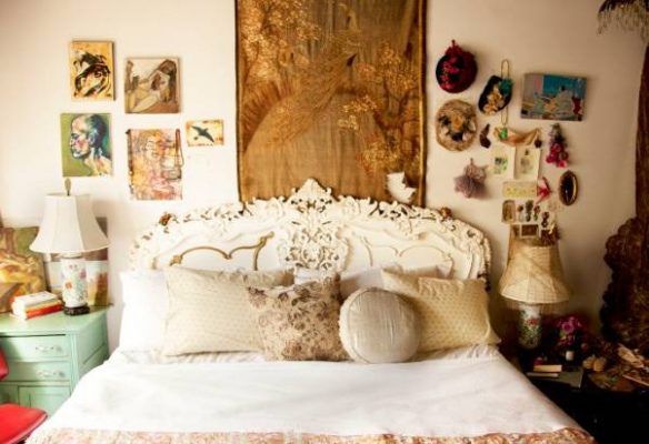 Un dormitorio de inspiración vintage con una cabecera blanca ornamentada, una variedad de almohadas decorativas y objetos y arte ecléctico en las paredes colocados sobre la cama.