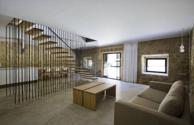 Espacio interior moderno caracterizado por una escalera con balaustres metálicos verticales, suelo de hormigón, paredes de piedra, un sofá beige y muebles minimalistas con grandes ventanales que ofrecen luz natural al estilo Decoración de casa.
