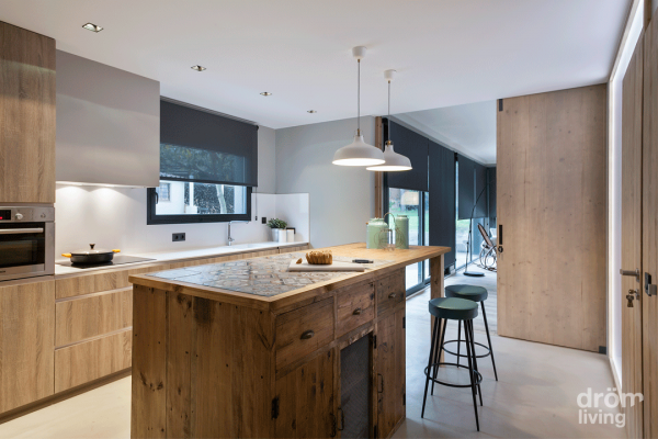 Interior de cocina moderno con una isla central con un frente de madera de estilo vintage, taburetes de bar, luces colgantes, electrodomésticos integrados y una paleta de colores suaves y neutros que resume la complicidad de Dröm Living.