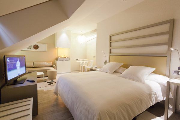 Una moderna habitación de Hotel&Spa con una cama grande con sábanas blancas, un televisor de pantalla plana con una imagen, muebles elegantes, iluminación ambiental y un diseño de techo inclinado que añade un toque extra.
