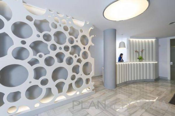 Una moderna área de recepción que presenta una partición decorativa perforada con formas circulares orgánicas, un elegante mostrador blanco y una persona contratada trabajando detrás del