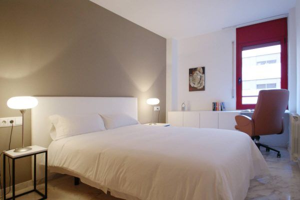 Una habitación minimalista orientada al alquiler, con una cama blanca cuidadosamente hecha, dos lámparas de mesilla, un escritorio con una silla,