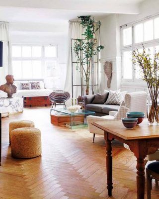 Una sala de estar luminosa y acogedora con pisos de madera, un sofá gris, un sillón blanco y varias plantas junto a las ventanas, complementada con una mezcla de elementos decorativos rústicos y modernos para una casa.