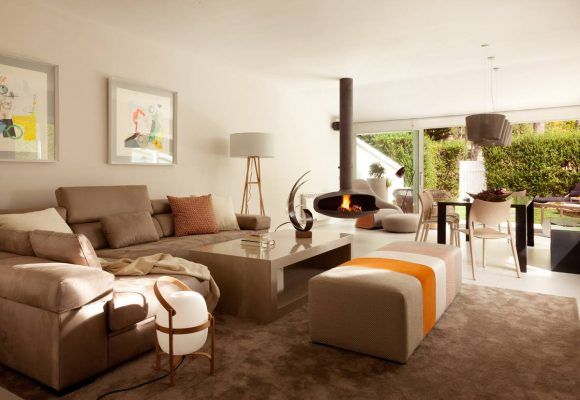 Una sala de estar moderna con un gran sofá seccional beige, una mesa de café central, una otomana naranja y una chimenea independiente, con obras de arte en las paredes de la Reforma de casa en