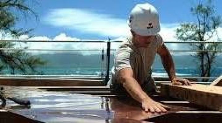 Una persona que lleva una gorra está trabajando en la estructura de una casa de madera al aire libre durante el verano, con un cielo azul claro y el océano al fondo.