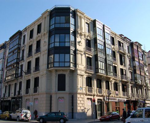 Una vista de la calle de un edificio de esquina con detalles arquitectónicos clásicos y elementos modernos, flanqueado por edificios tradicionales en hilera bajo un cielo azul claro, insinuando obras en curso.