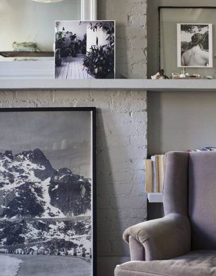 Un rincón acogedor de una habitación presentada en nuestro recorrido por la casa, que muestra un sillón gris junto a una colección de fotografías enmarcadas y obras de arte apoyadas contra una pared de ladrillos grises, con una pequeña estantería escondida al lado del