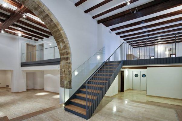 Un espacio interior moderno caracterizado por una escalera con peldaños de madera y barandilla metálica, un arco curvo rehabilitado y techos con vigas de madera vistas en el Palau del Ossets, Castellón.