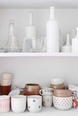 Una variedad de utensilios de cocina de cerámica y vidrio cuidadosamente dispuestos en dos estantes blancos para un recorrido luminoso por la casa.