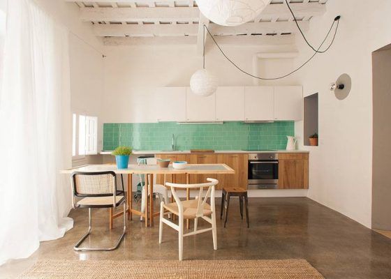 Una cocina moderna con paredes blancas, un protector contra salpicaduras de azulejos verdes, gabinetes de madera clara y un área de comedor con una mesa de madera y una combinación de sillas le espera en el recorrido por una casa en Barcelona.