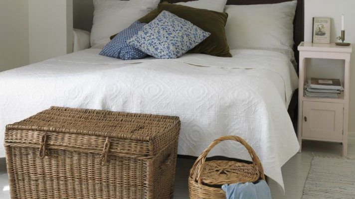 Un dormitorio con una cama bien hecha, un edredón blanco, almohadas variadas, un cesto para ropa de mimbre y otro pequeño canasto con tela.